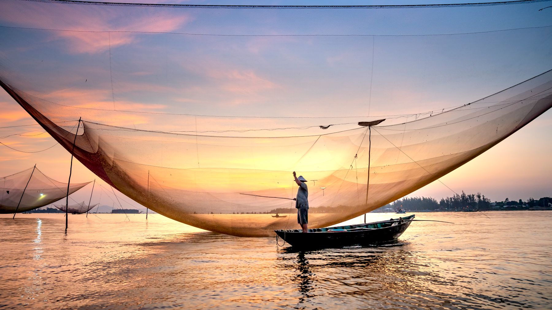 Một ngư dân quăng chài lúc hoàng hôn trên vùng nước ở Hội An, Việt Nam, thể hiện các phương thức đánh bắt truyền thống.
