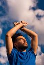 From below of tranquil young bearded male in blue t shirt with gauged ears enjoying atmosphere with raised hands and closed eyes