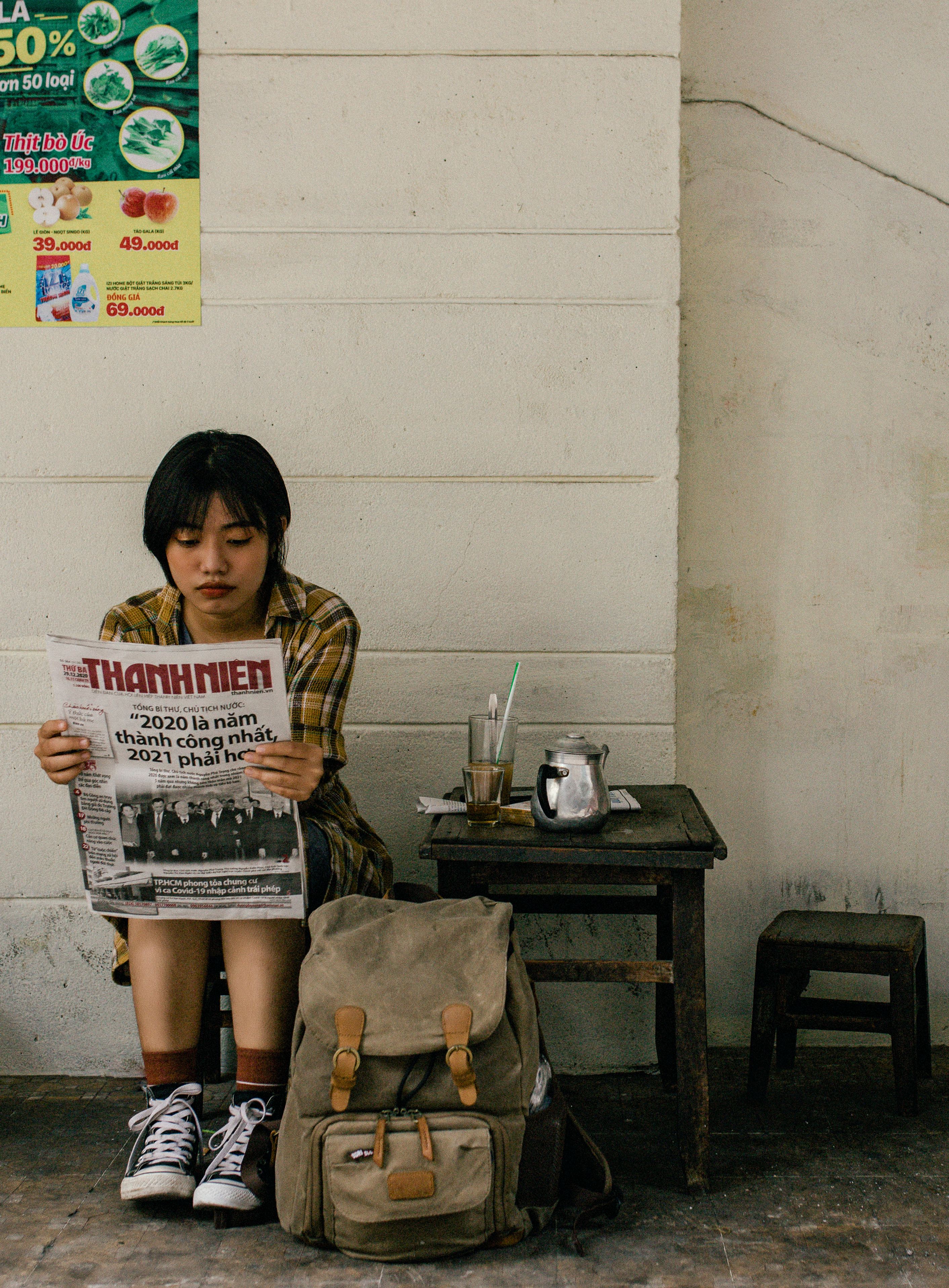 Young Asian woman in casual clothes and sneakers sitting on small stool and reading newspaper near backpack and stool with glass of beverage