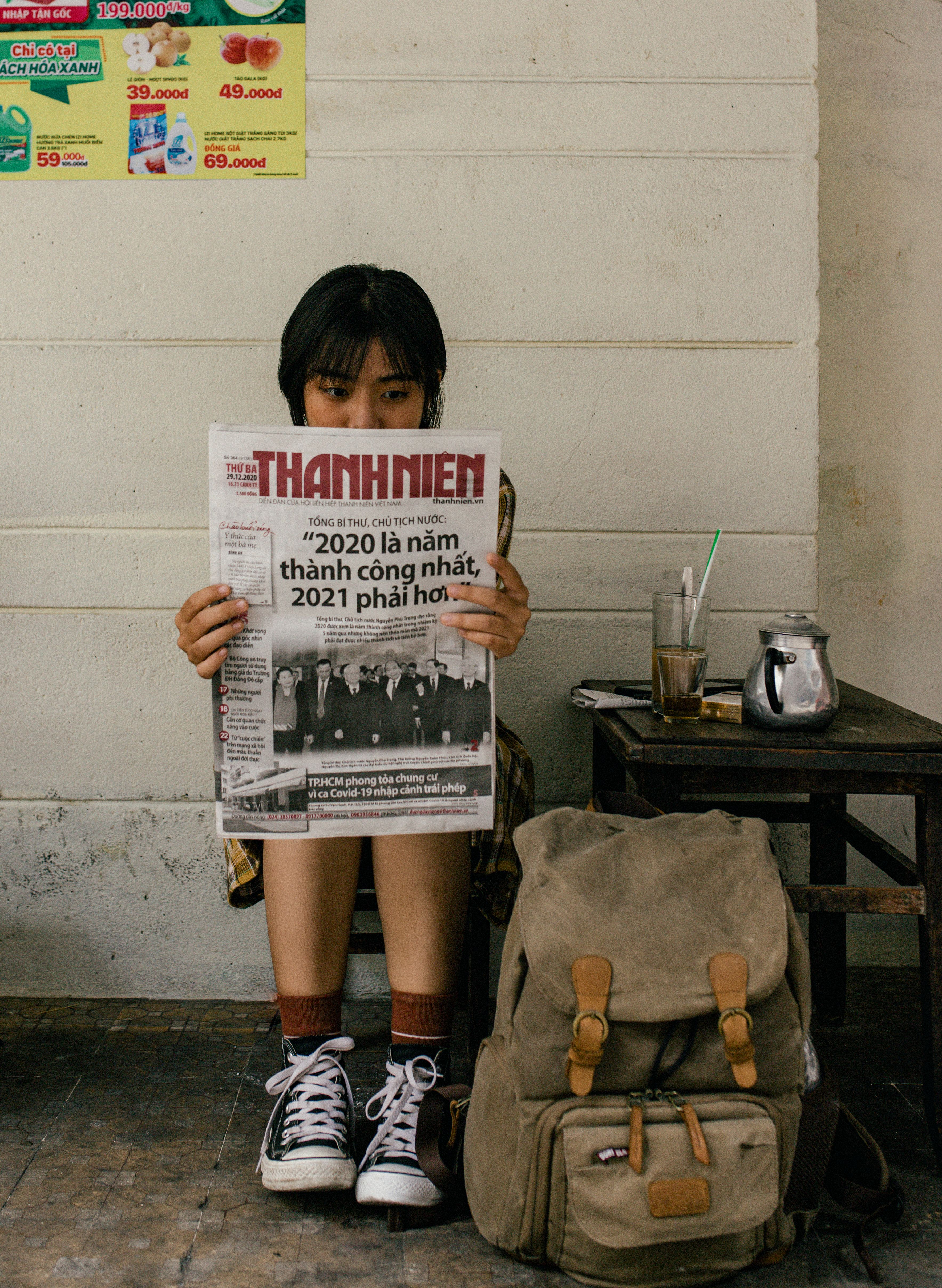 Young Asian woman reading newspaper beside stool with glass of beverage and backpack