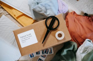 Top view of tape with scissors on carton box with exact address and name among clothes