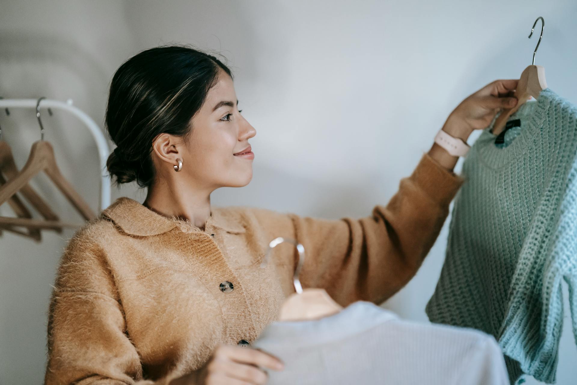 Cheerful young female in stylish cardigan smiling and choosing clothes on hangers in wardrobe