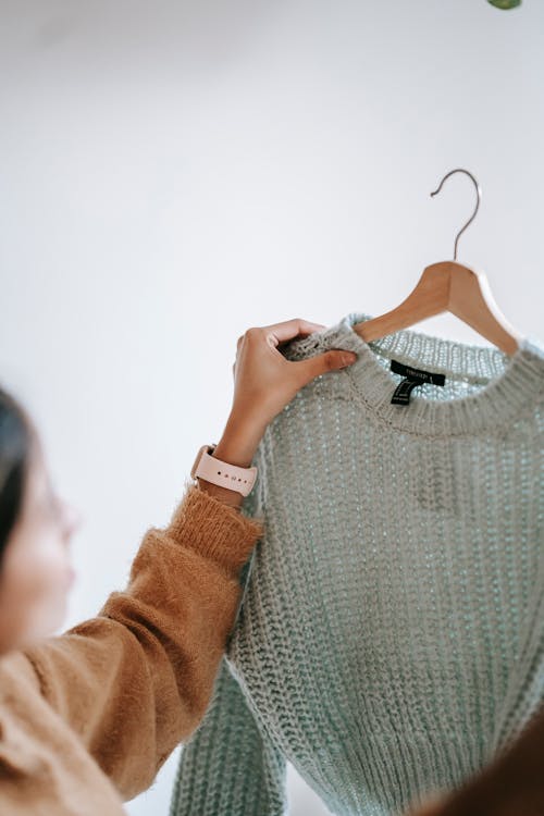 Crop anonymous female designer in casual clothes demonstrating new sweater on hanger while standing in room