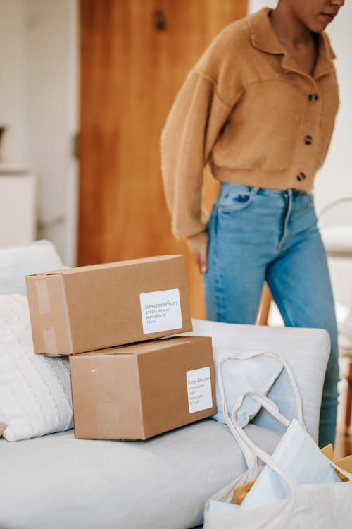 Crop unrecognizable female standing near couch with carton boxes and shopping bag while preparing for delivering