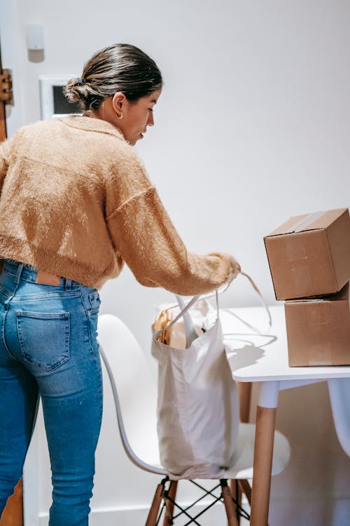 Back view of young female packing shopping bag with carton boxes while standing near table