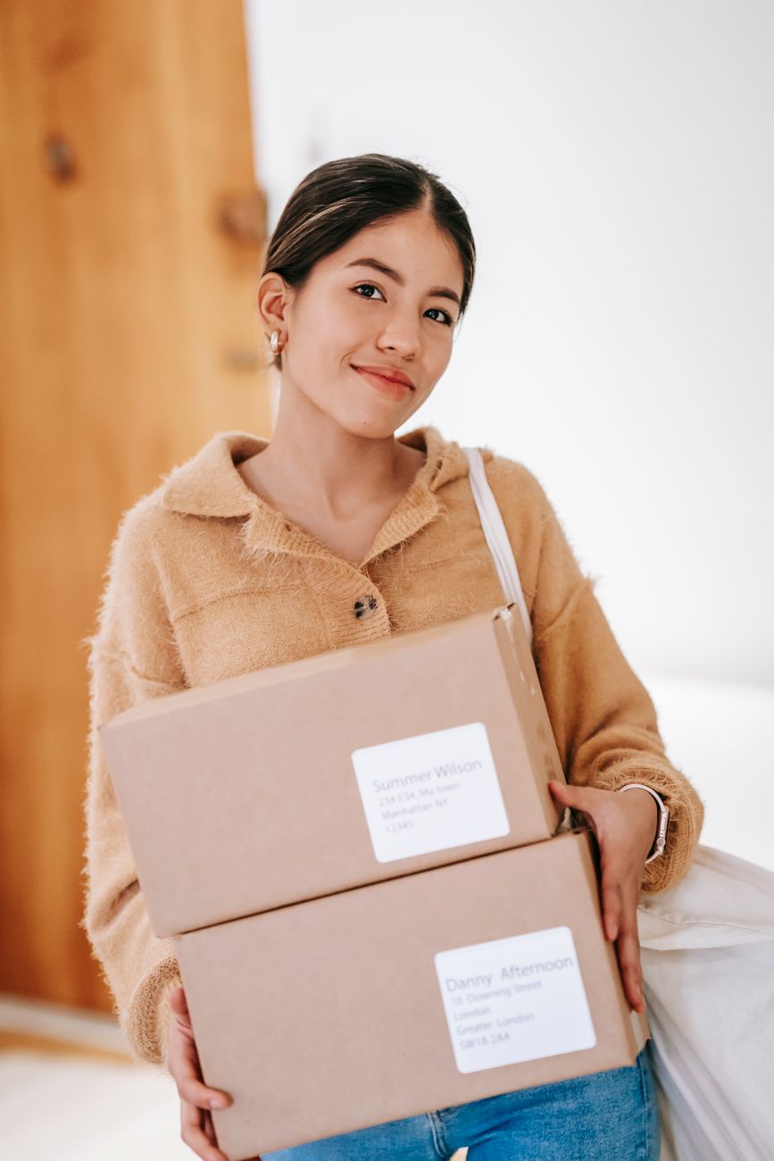 Positive ethnic female with heavy shopping bag and cardboard boxes standing in apartment and looking at camera
