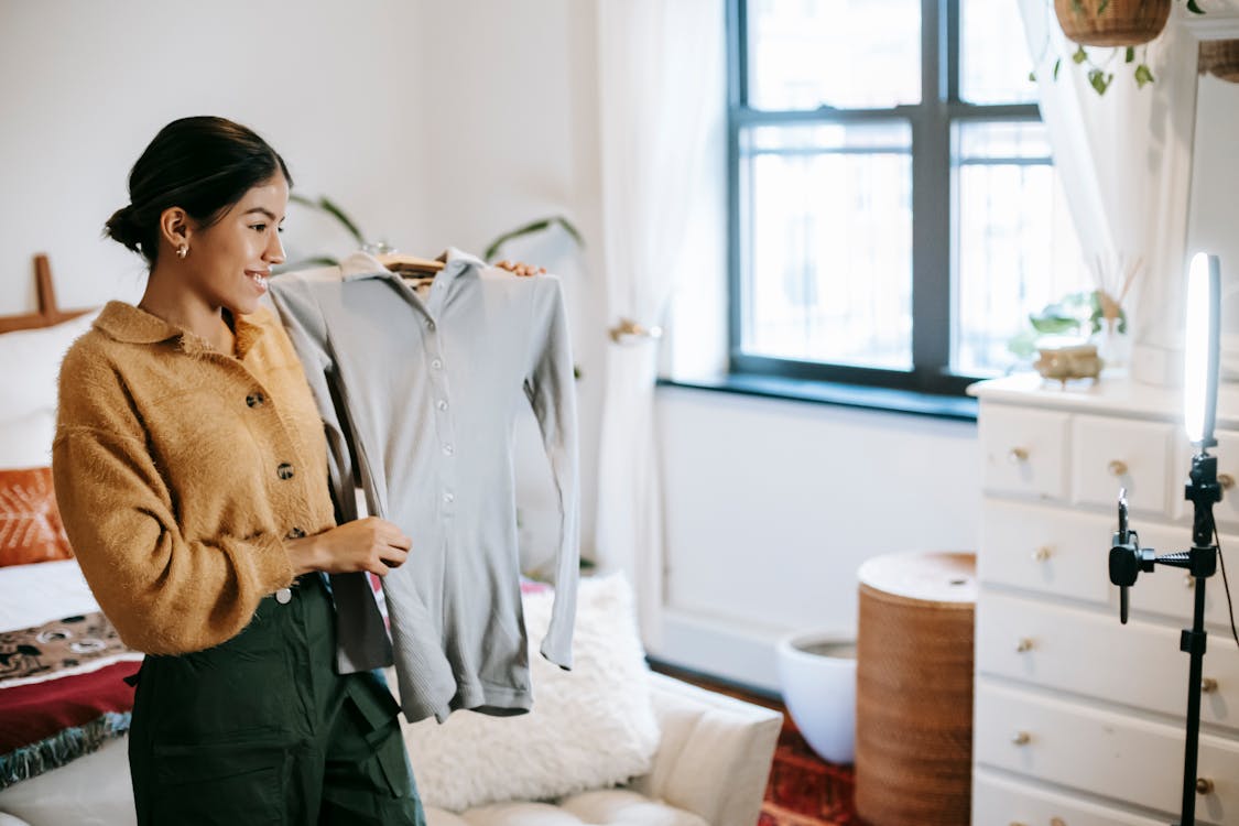 Free Positive ethnic female blogger demonstrating jacket on hanger during live stream on mobile phone placed on shining lamp Stock Photo | influencer marketing