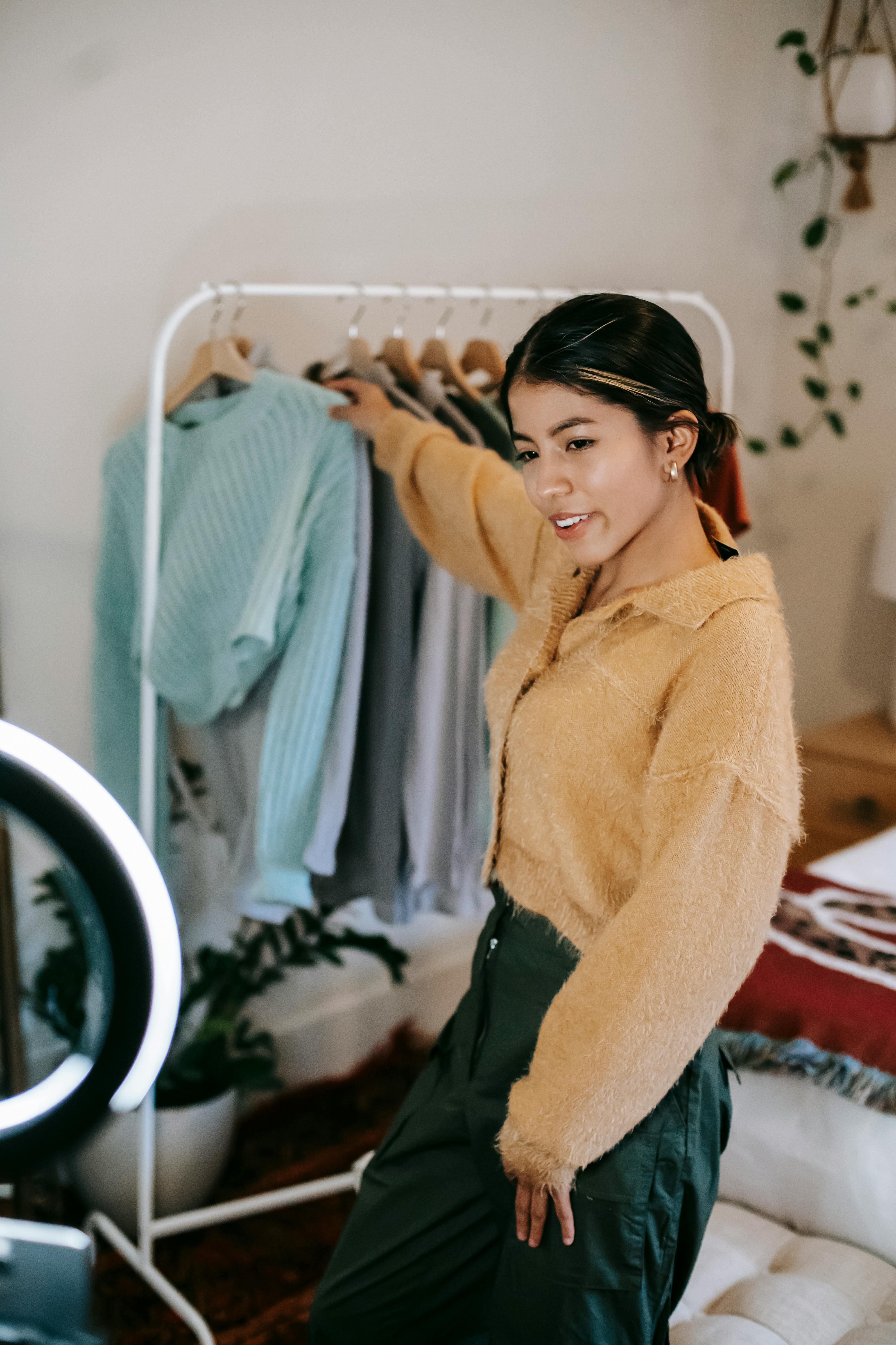 trendy smiling woman taking clothes from rail while shooting video