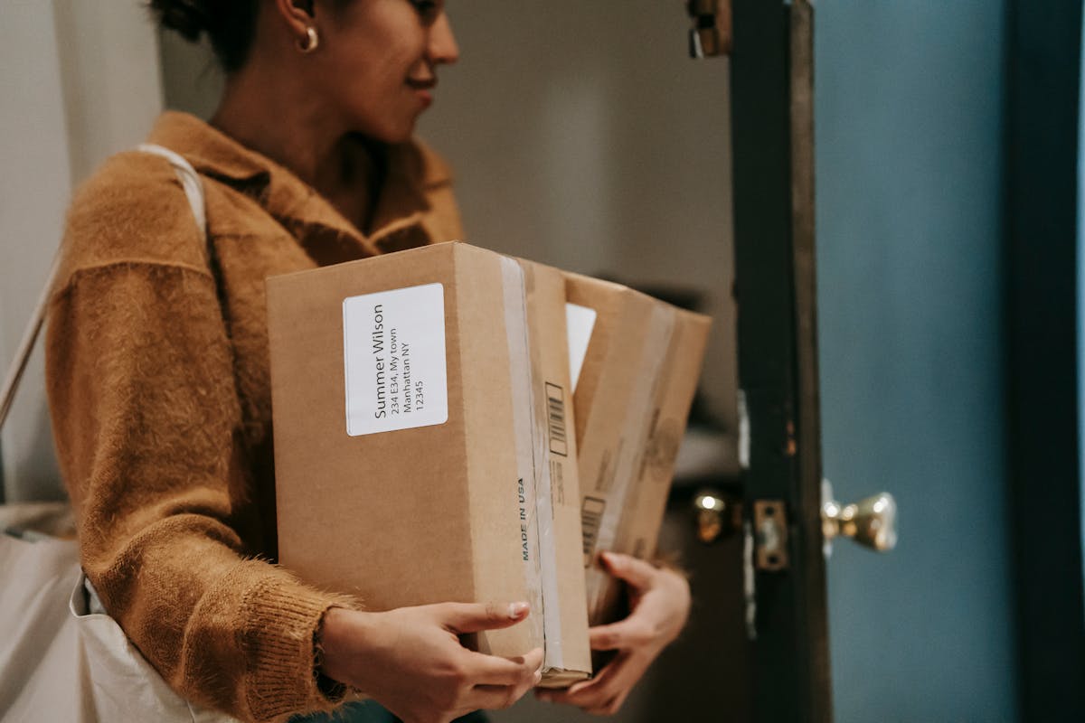 Crop ethnic female walking into open door of apartment with carton boxes with goods from delivery