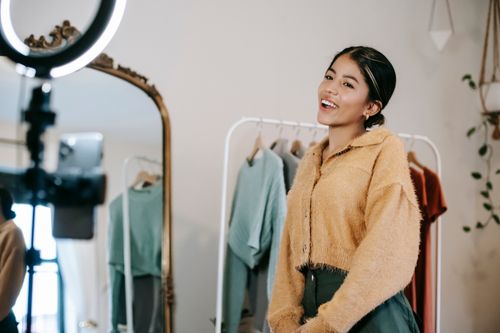 Free Positive young ethnic female blogger standing in wardrobe and using phone with ring light while filming vlog near mirror and trendy clothes Stock Photo
