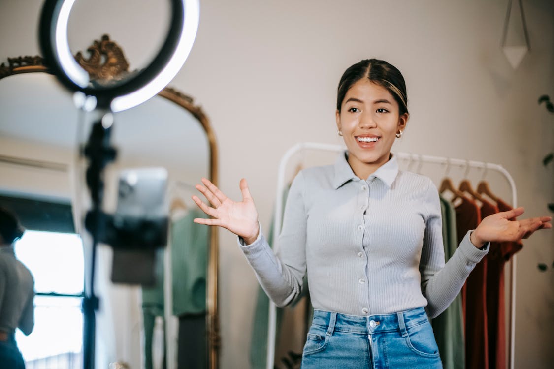 Free Happy young ethnic female blogger in blouse and jeans standing in wardrobe and using phone with ring light while filming vlog about trendy clothes near mirror Stock Photo