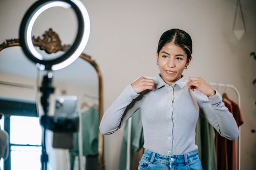 Free Young ethnic female blogger in blouse and jeans standing in wardrobe and using cellphone with ring light while taking selfie and shooting video about trendy clothes near mirror Stock Photo