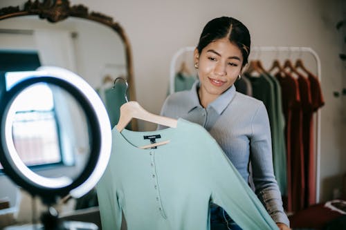 Smiling young ethnic female in trendy clothes standing in room near wardrobe and mirror and demonstrating blouse on hanger on ring lamp with cellphone