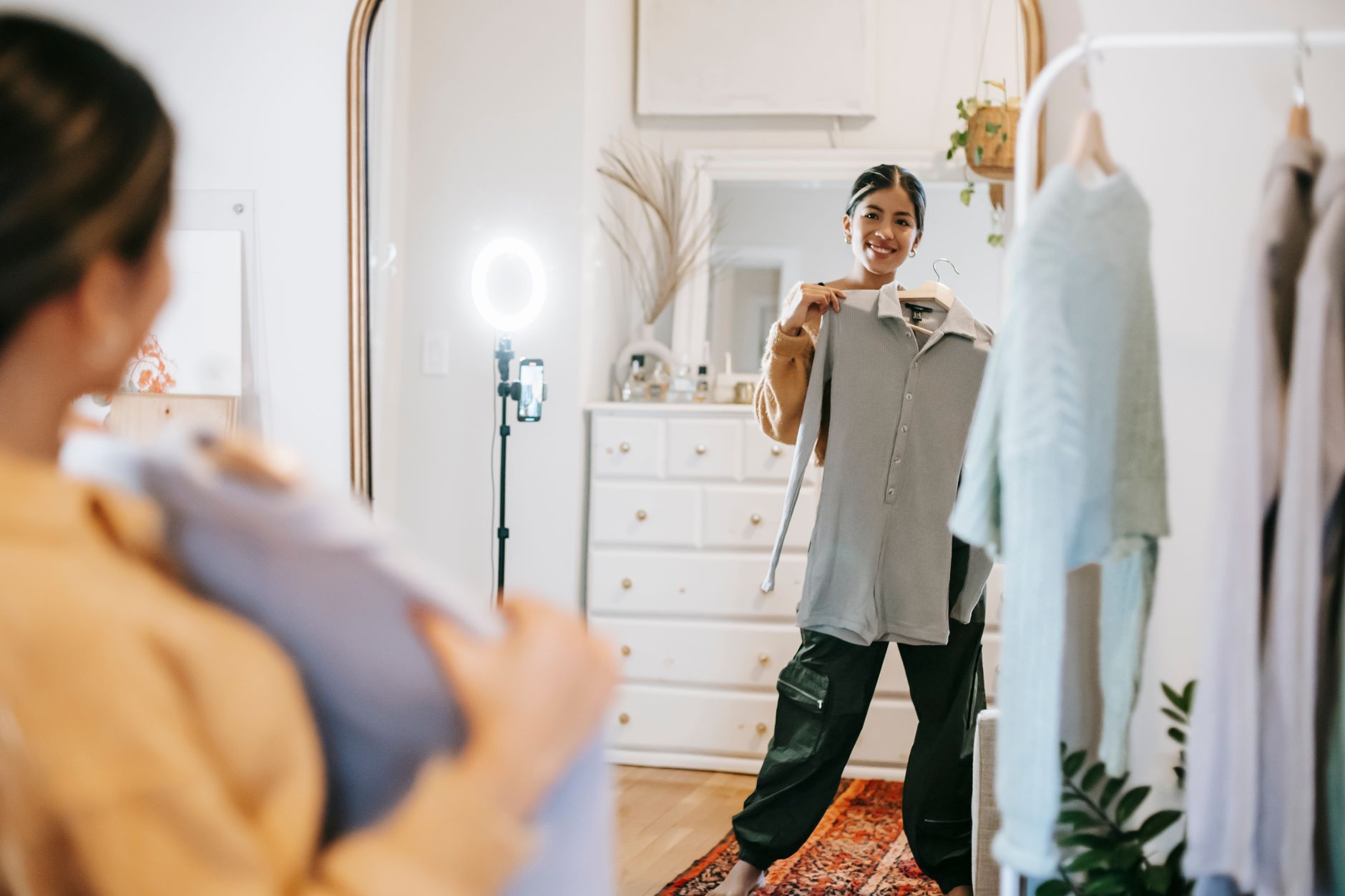 How to Film Q&A Videos on Your Phone: A Practical Beginner Guide Smiling young ethnic female blogger standing in room near wardrobe while demonstrating blouse on hanger and looking in mirror and filming video on cellphone with ring lamp