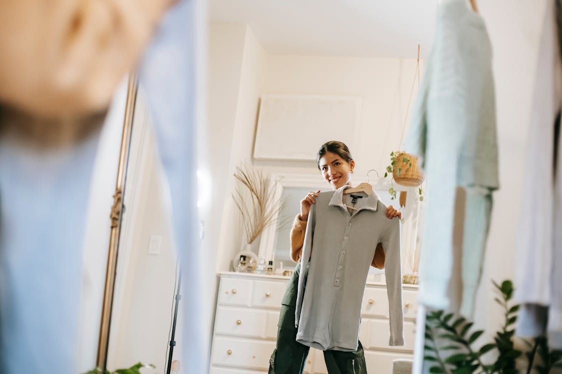 Smiling young ethnic woman in casual outfit demonstrating blouse on hanger while selecting clothes and looking in mirror in room near cabinet and wardrobe at bright home