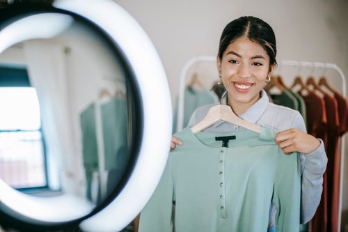 Free Smiling young ethnic woman in casual outfit demonstrating blue blouse on hanger while selecting clothes near ring lamp and mirror near wardrobe in bright room at home Stock Photo