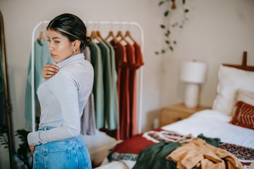 Side view of confident ethnic woman in trendy outfit demonstrating outfit in bedroom near bed and wardrobe near mirror at home