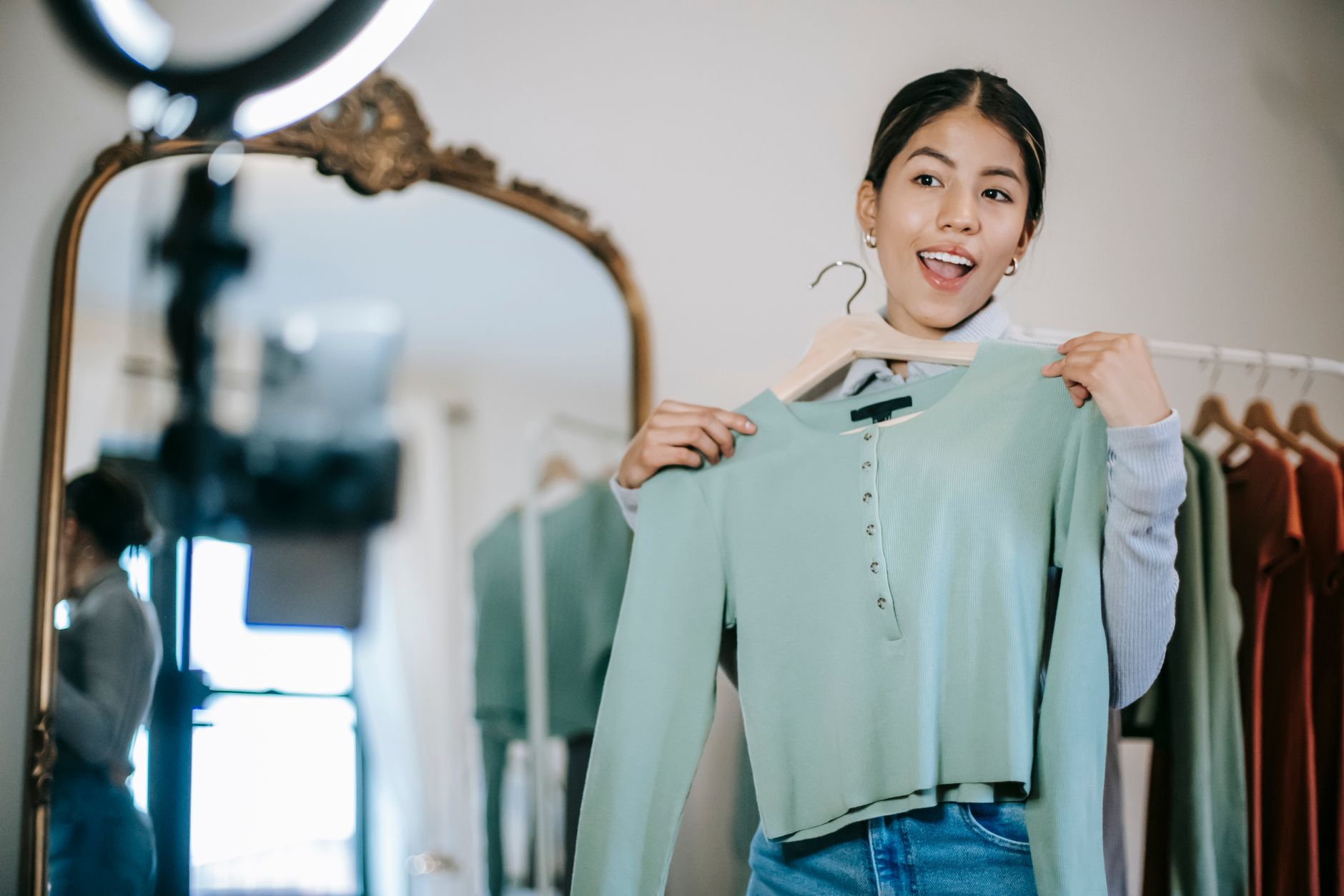 Smiling young ethnic female blogger standing in room near wardrobe and mirror while demonstrating blouse on hanger and filming vlog on mobile with ring lamp