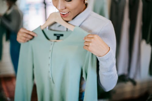 Crop unrecognizable smiling woman in trendy outfit choosing clothes in light shop with blue blouse on hanger in hands