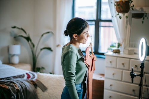 Side view of smiling faceless young ethnic female blogger standing in bedroom and demonstrating pink dress on hanger while filming video on cellphone with ring lamp near cupboard and window