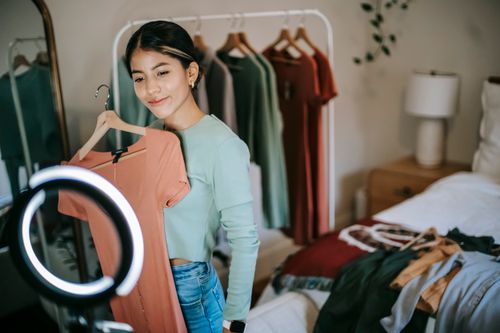 Free Happy young ethnic female blogger standing in bedroom and demonstrating pink dress on hanger while filming video on smartphone with ring lamp near bed and mirror near wardrobe Stock Photo