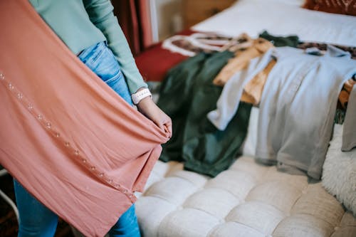 Crop anonymous woman in casual clothes demonstrating pink dress while selecting clothes while standing in bright bedroom near couch and bed with wardrobe at home