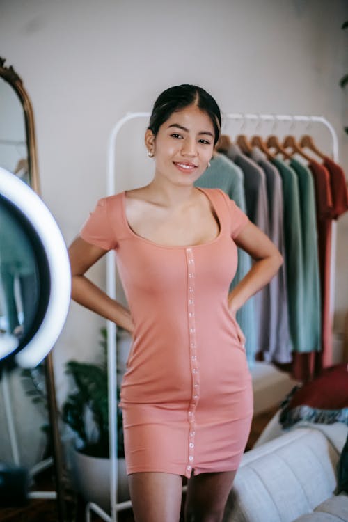 Free Smiling young ethnic female blogger shooting video on phone with ring lamp while demonstrating pink dress near mirror and wardrobe near bed with sofa near potted plant in bedroom Stock Photo
