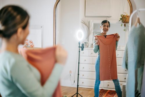 Free Smiling young ethnic female blogger in trendy outfit standing in room and demonstrating dress in hands while looking at mirror and shooting video on phone with ring lamp near cabinet Stock Photo