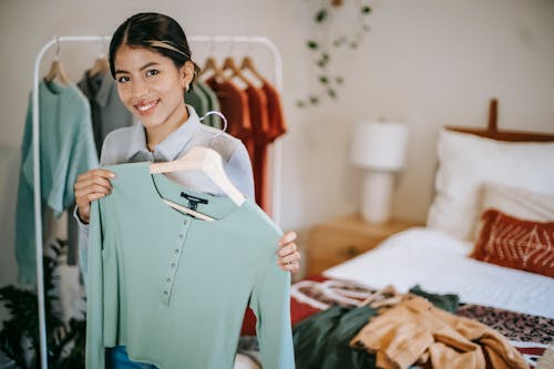 Free Positive young ethnic woman in trendy outfit choosing and demonstrating clothes in bedroom at light home near bed and wardrobe and looking at camera Stock Photo