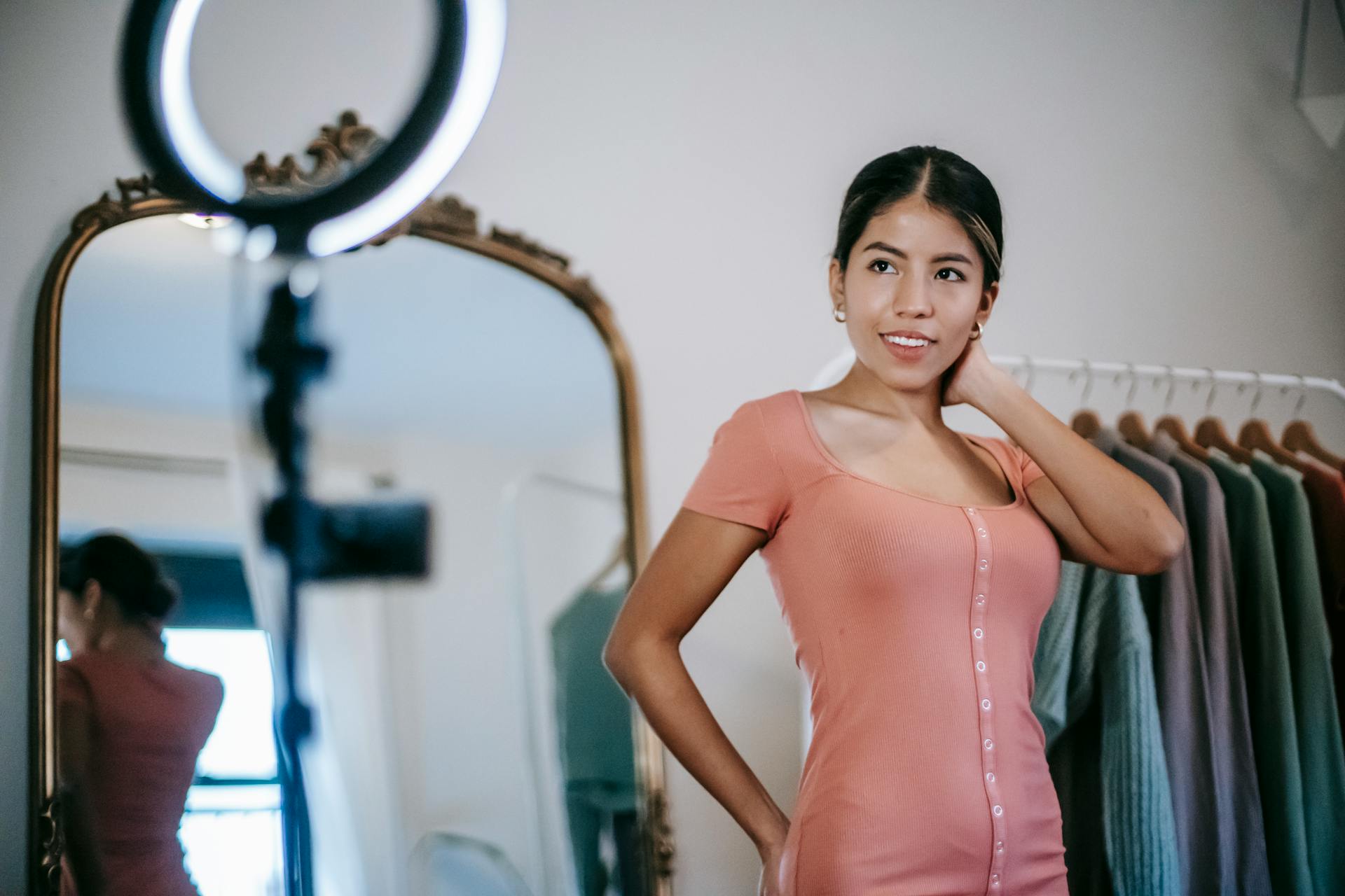 Confident young ethnic female blogger demonstrating pink dress while standing in room and filming video on phone with ring lamp near mirror and wardrobe