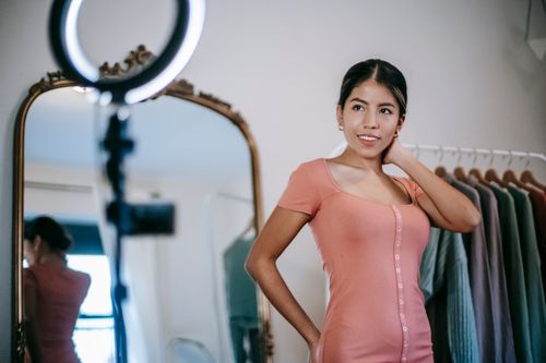Free Confident young ethnic female blogger demonstrating pink dress while standing in room and filming video on phone with ring lamp near mirror and wardrobe Stock Photo