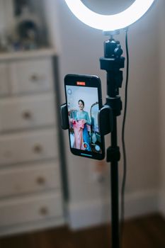 Faceless female blogger in trendy outfit standing in room and shooting video on phone with ring lamp while demonstrating pink dress in hands near cabinet