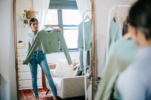 Free Serious young ethnic woman in trendy outfit choosing and demonstrating blouse on hanger in room near couch and cabinet at light home near wardrobe and looking in mirror Stock Photo