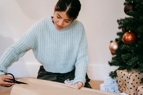 Crop ethnic craftswoman cutting wrapping paper on Christmas day indoors