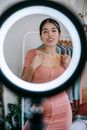 Young feminine ethnic female in dress touching chest while looking at camera behind light ring in house room