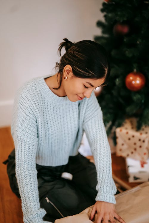 Sincere young ethnic female in warm clothes with wrapping paper preparing for New Year holiday in house