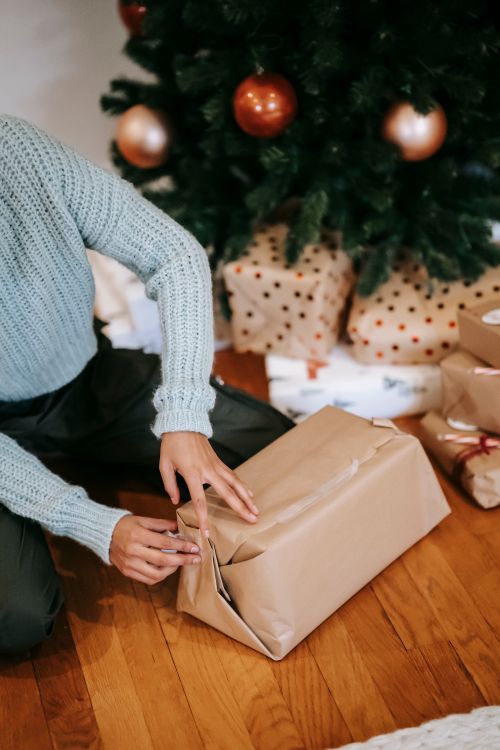 Free Crop anonymous female wrapping present box on floor against Christmas tree with baubles at home Stock Photo
