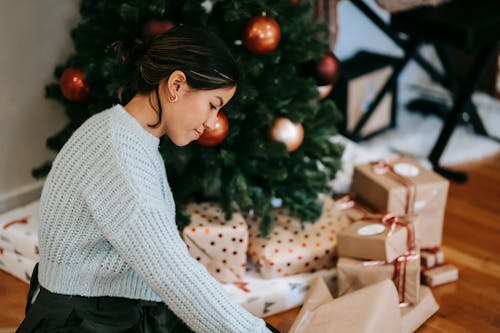Free Side view of young ethnic female sitting on parquet with heap of gift boxes during New Year holiday and looking down at home Stock Photo