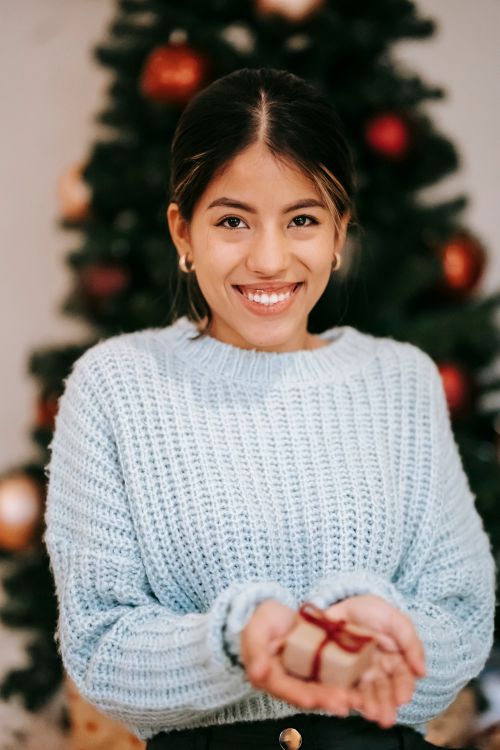Free Cheerful ethnic female in knitted sweater with small present box looking at camera during Christmas holiday at home Stock Photo