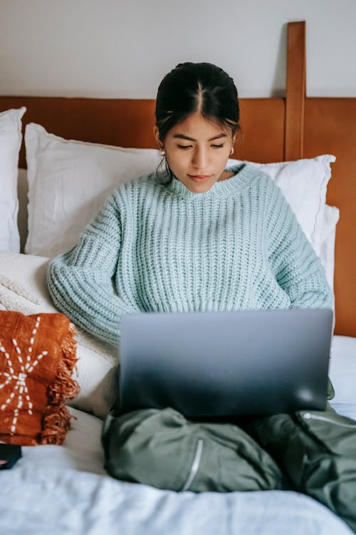 Young attentive ethnic female surfing internet on portable computer while sitting on bed at home