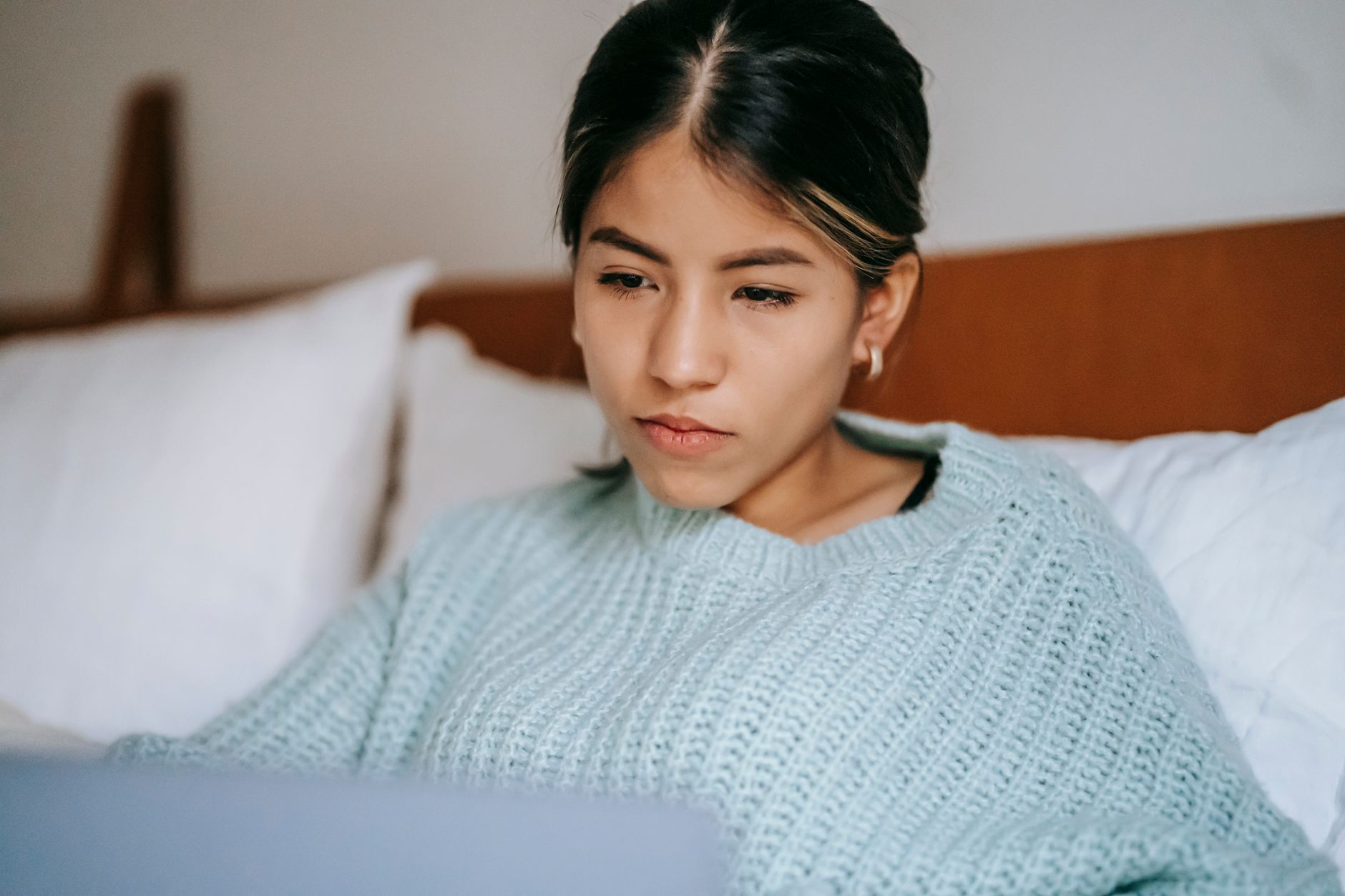 Young woman in a sea-green sweater sitting on a bed, looking at her laptop screen