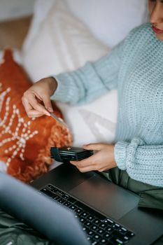 Crop female buyer with debit card and netbook shopping online on bed in house