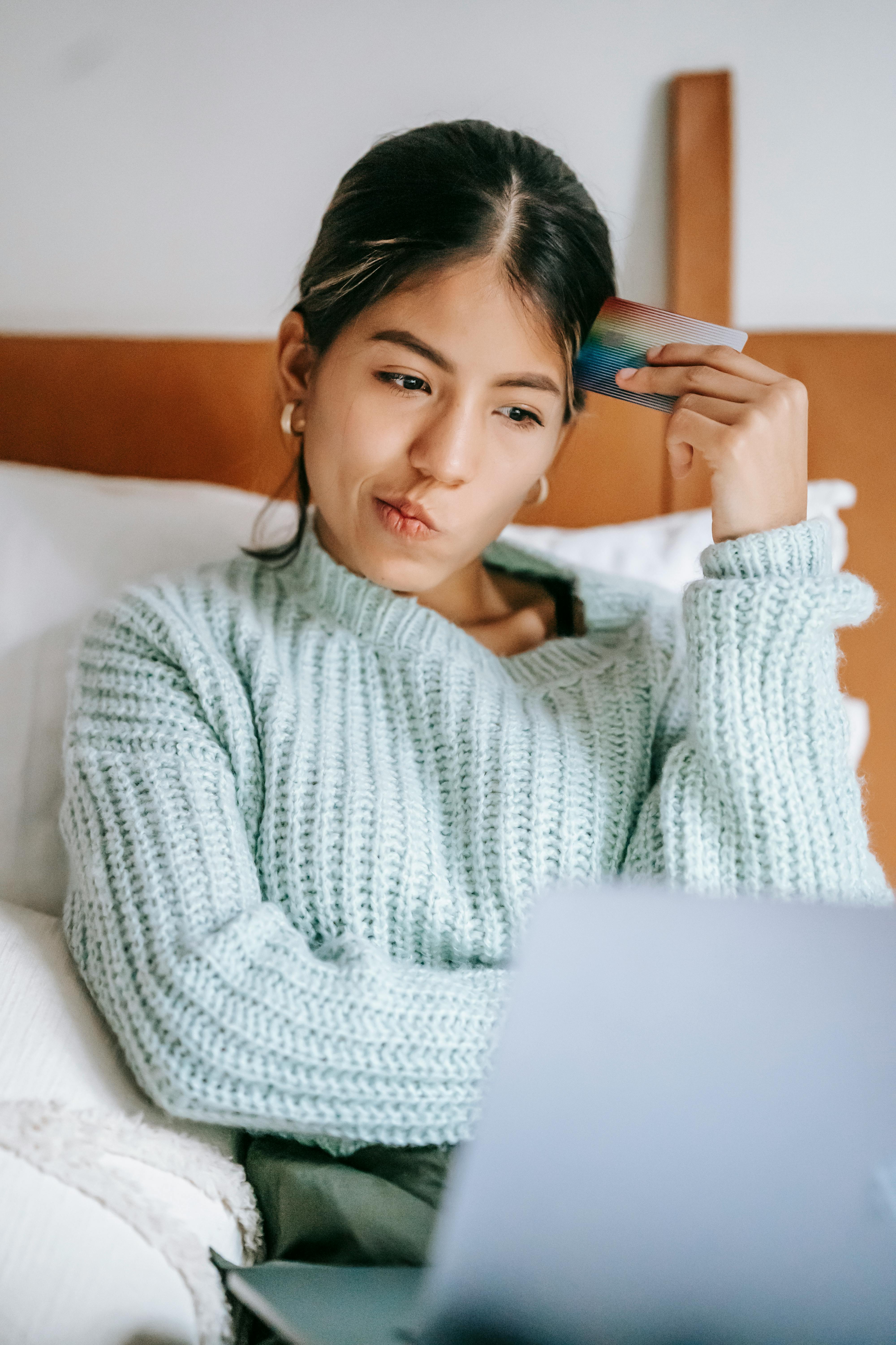 pensive ethnic shopper with debit card and laptop on bed