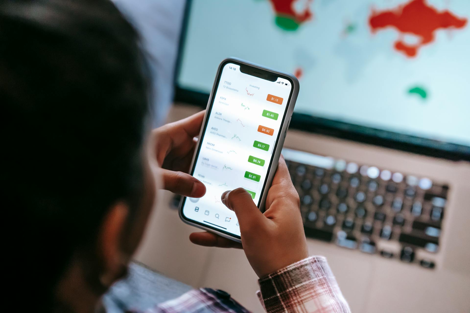 A person checks stock market graphs on a smartphone with a laptop in the background.