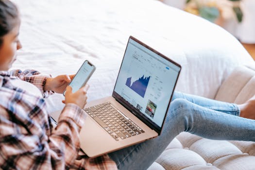 A woman using a smartphone and laptop to analyze stock market data from home.