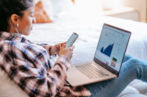 Free Side view of crop ethnic female dealer using trading app on cellphone against netbook with graph on screen in house Stock Photo