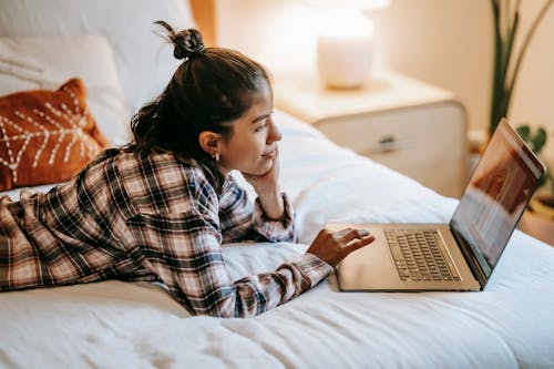 Side view of concentrated ethnic woman freelancer with dark hair in casual clothes lying on comfortable bed and browsing laptop at home