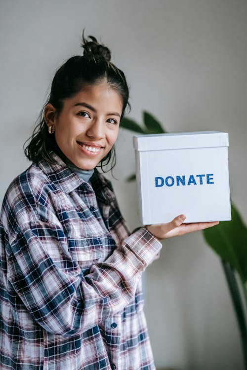 Happy ethnic lady in checkered shirt and bun looking at camera and smiling while demonstrating donation box on background with potted plant near white wall