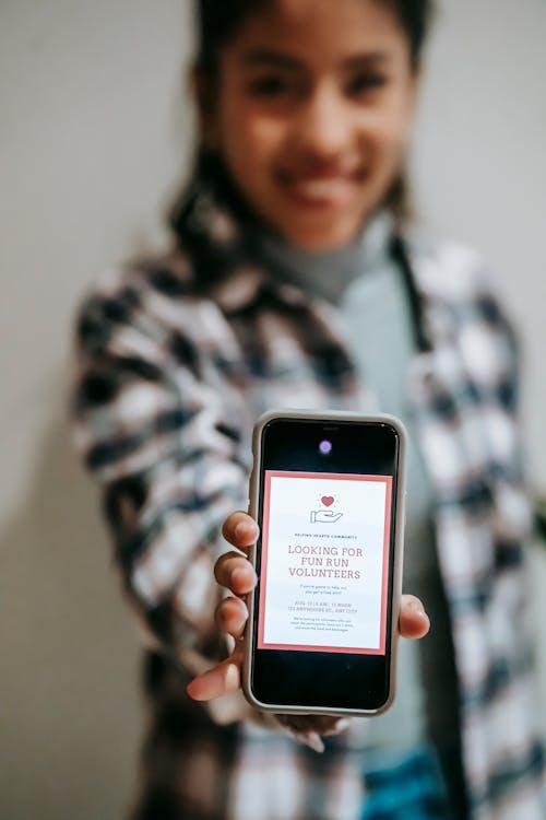 Crop blurred ethnic happy lady in checkered shirt demonstrating app with invitation to be volunteer on screen of smartphone on gray background