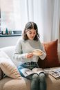 Focused lady in casual clothes sitting with cup of hot drink and saucer on comfortable sofa and reading magazines in cozy living room